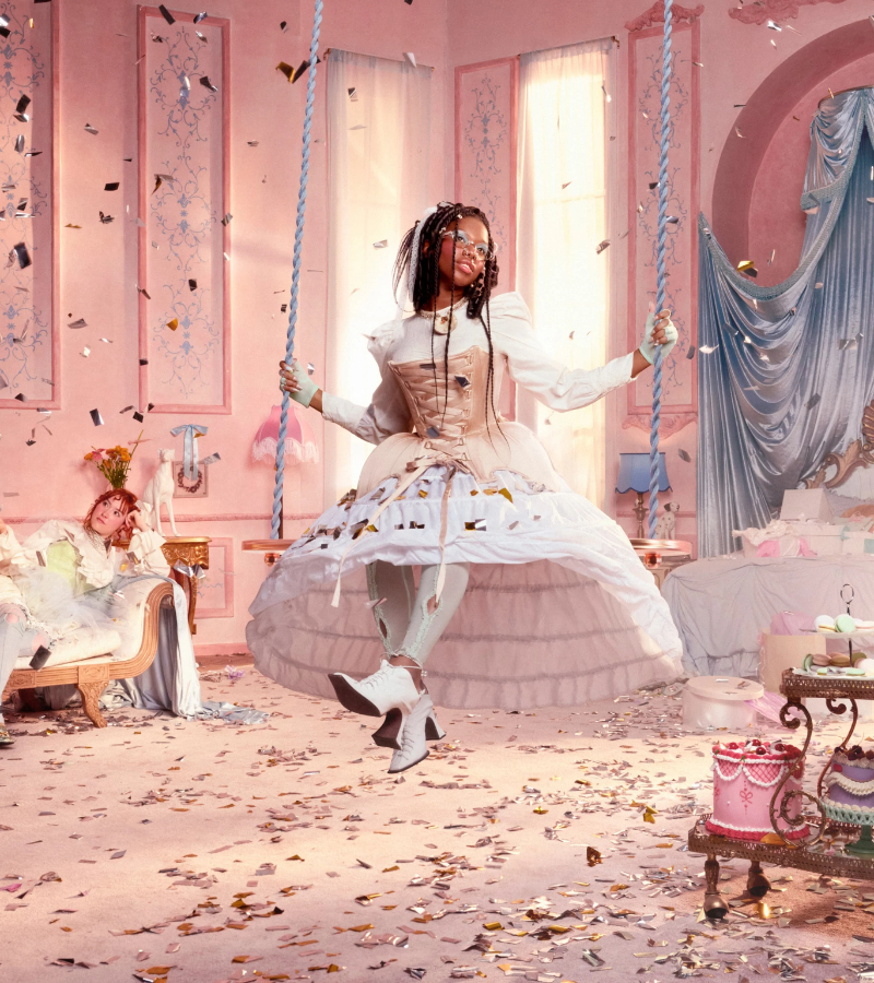 Rococo Revival scene of a woman wearing a beige corset and white hoop skirt sitting on rope swing as confetti falls.