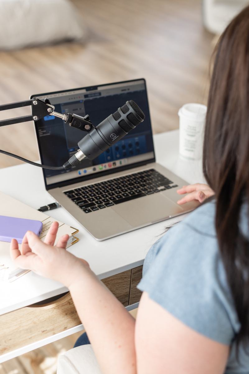 Woman recording a podcast beside a laptop and microphone while discussing marketing ideas, content planning, and business strategy at a bright home office desk.