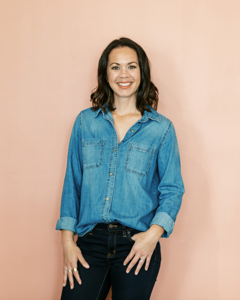 Tayler Cusick Holman, co-founder of Enji Marketing Software smiling in a denim shirt and dark jeans stands confidently against a pink background, embodying approachable marketing planning expertise.