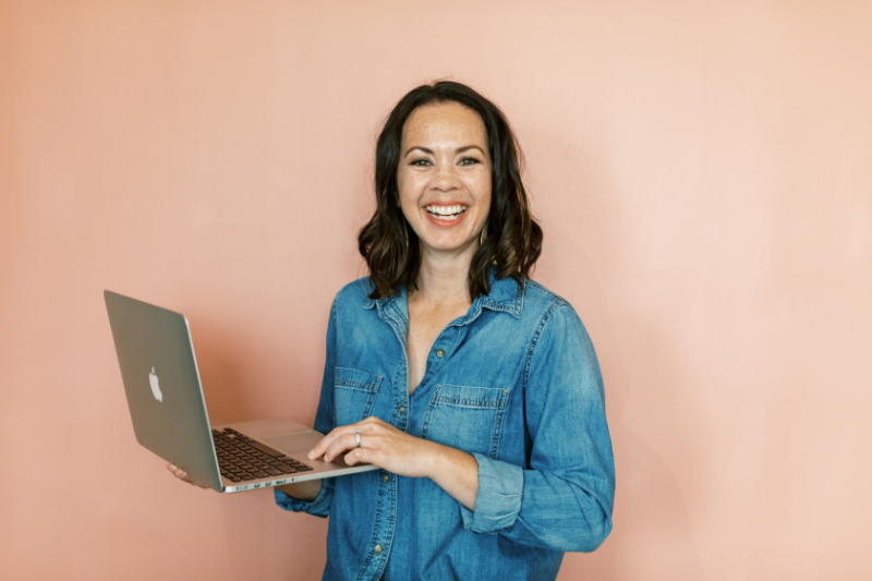 Tayler Cusick-Hollman, co-founder of Enji Marketing Software, smiling while holding a laptop against a pink wall, representing how small business owners may be sabotaging your marketing without realizing it.