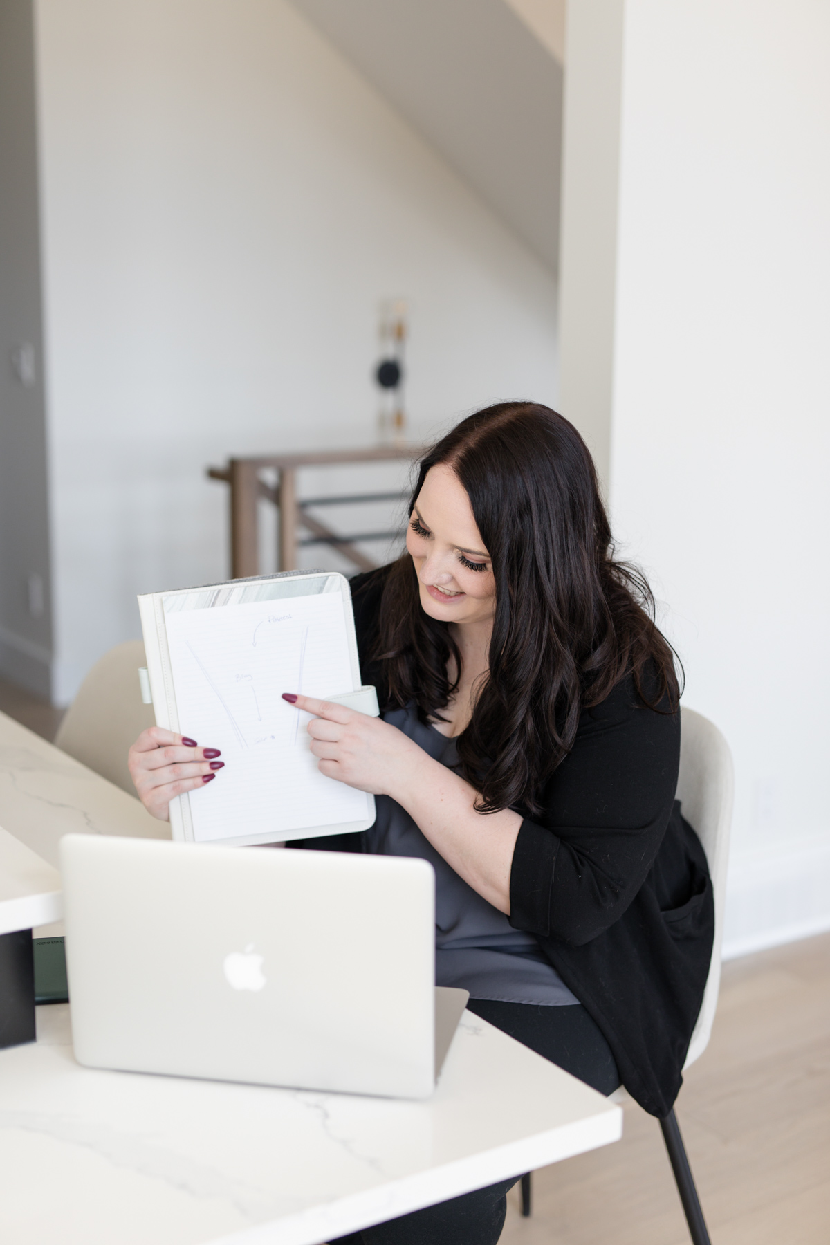 A smiling female entrepreneur sits at her desk, pointing to notes in her planner while presenting to her laptop. She shares insights and strategies to help business owners sound like an expert and communicate with confidence.